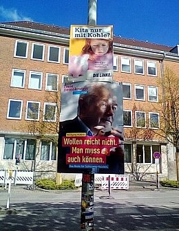 Es hängen zwei Wahlplakte übereinander an einem Laternenmast: Oben ein Plakat der Partei DIE LINKE mit dem Slogan "Kita nur mit Kohle?", unten ein größeres Plakat der FDP. Auf dem Plakat der FDP ist Wolfgang Kubicki zu sehen, darunter der Slogan "Wollen reicht nicht. Man muss auch können."